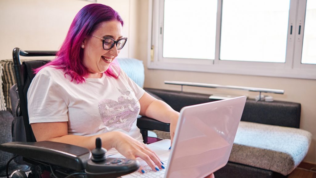 Smiling woman with purple hair in a wheelchair using a white laptop.