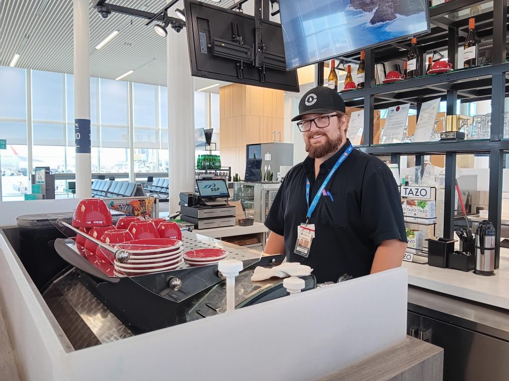 Caleb smiling behind the counter at Spinnakers on the Fly in Victoria International Airport, wearing a black uniform, cap, glasses, and an airport ID lanyard.