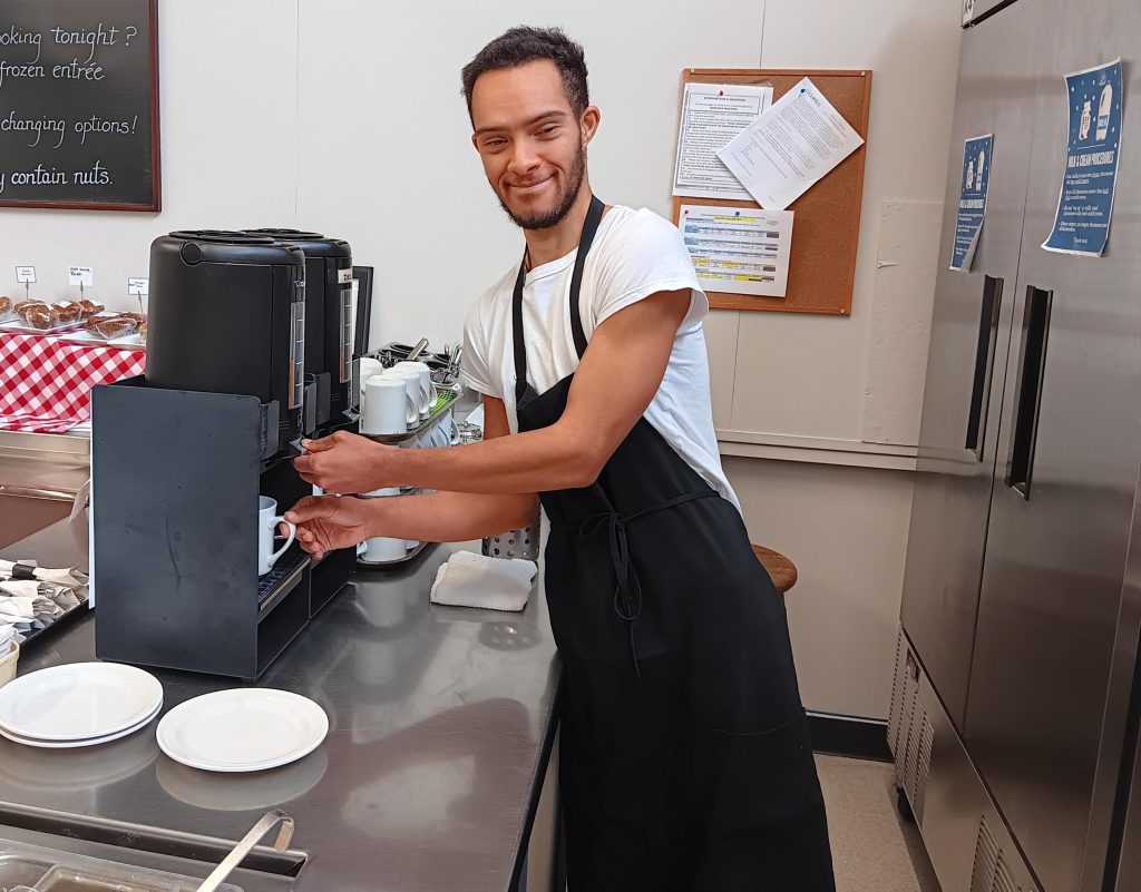 Smiling young man in a white t-shirt and black apron pouring coffee from a dispenser into a white ceramic mug at a community café.